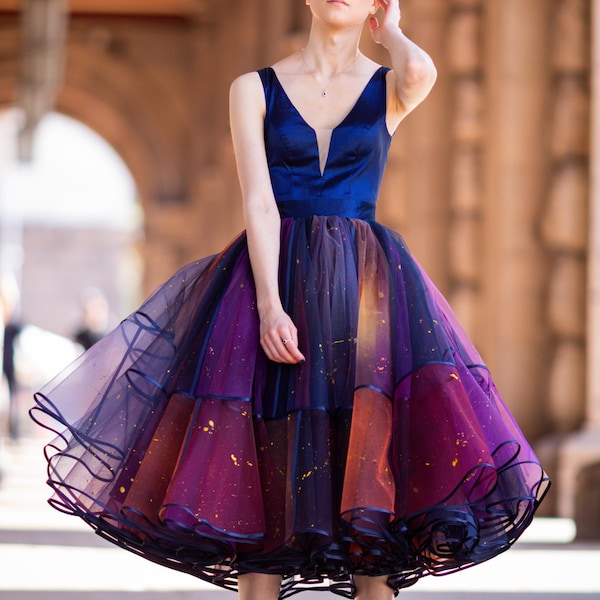 May include: A knee-length dress featuring a dark blue satin bodice with a deep V-neckline and a sheer mesh insert. The voluminous skirt is crafted from layers of dark blue and purple tulle, accented with scattered gold paint splatters.