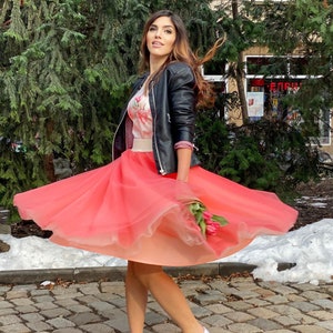 May include: A woman wearing a black leather jacket and a pink tulle skirt, holding a bouquet of pink flowers. She is standing on a cobblestone street with snow on the ground.