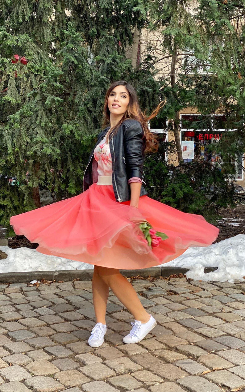 May include: A woman wearing a black leather jacket and a pink tulle skirt, holding a bouquet of pink flowers. She is standing on a cobblestone street with snow on the ground.