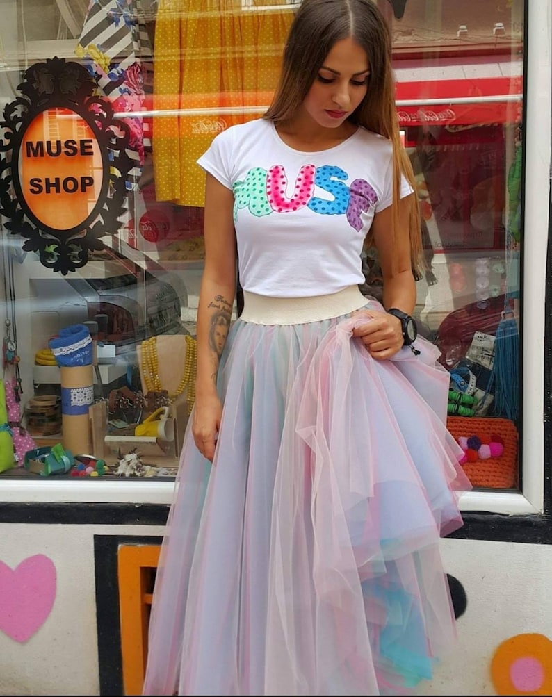 May include: A woman wearing a white t-shirt with the word "MUSE" in colourful letters and a pink tulle skirt with rainbow accents.