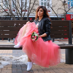 May include: A woman wearing a black leather jacket and a pink tulle skirt sits on a wooden bench. She is holding a bouquet of pink tulips.