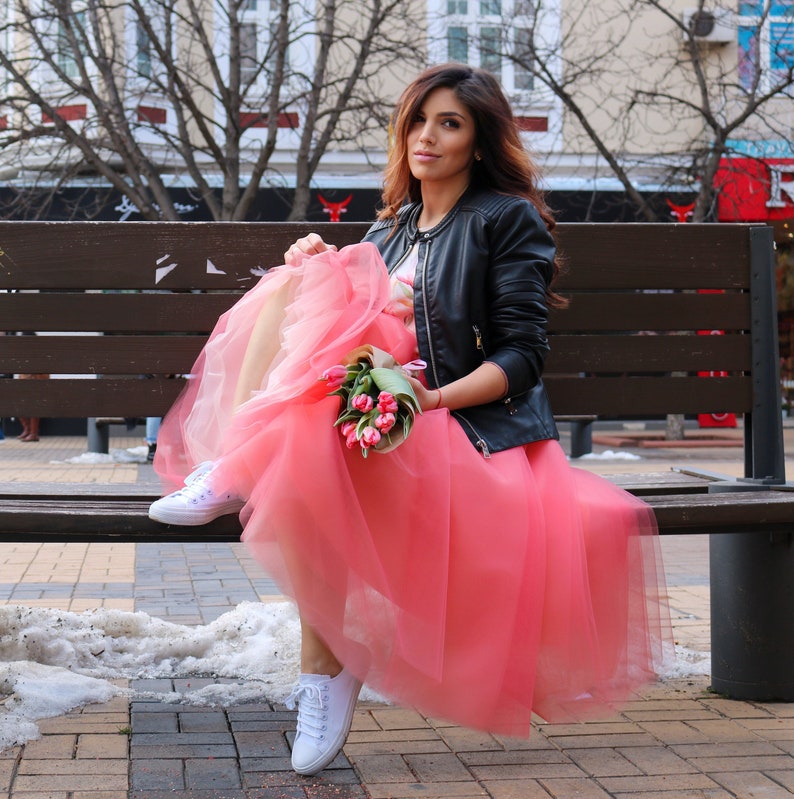 May include: A woman wearing a black leather jacket and a pink tulle skirt sits on a wooden bench. She is holding a bouquet of pink tulips.