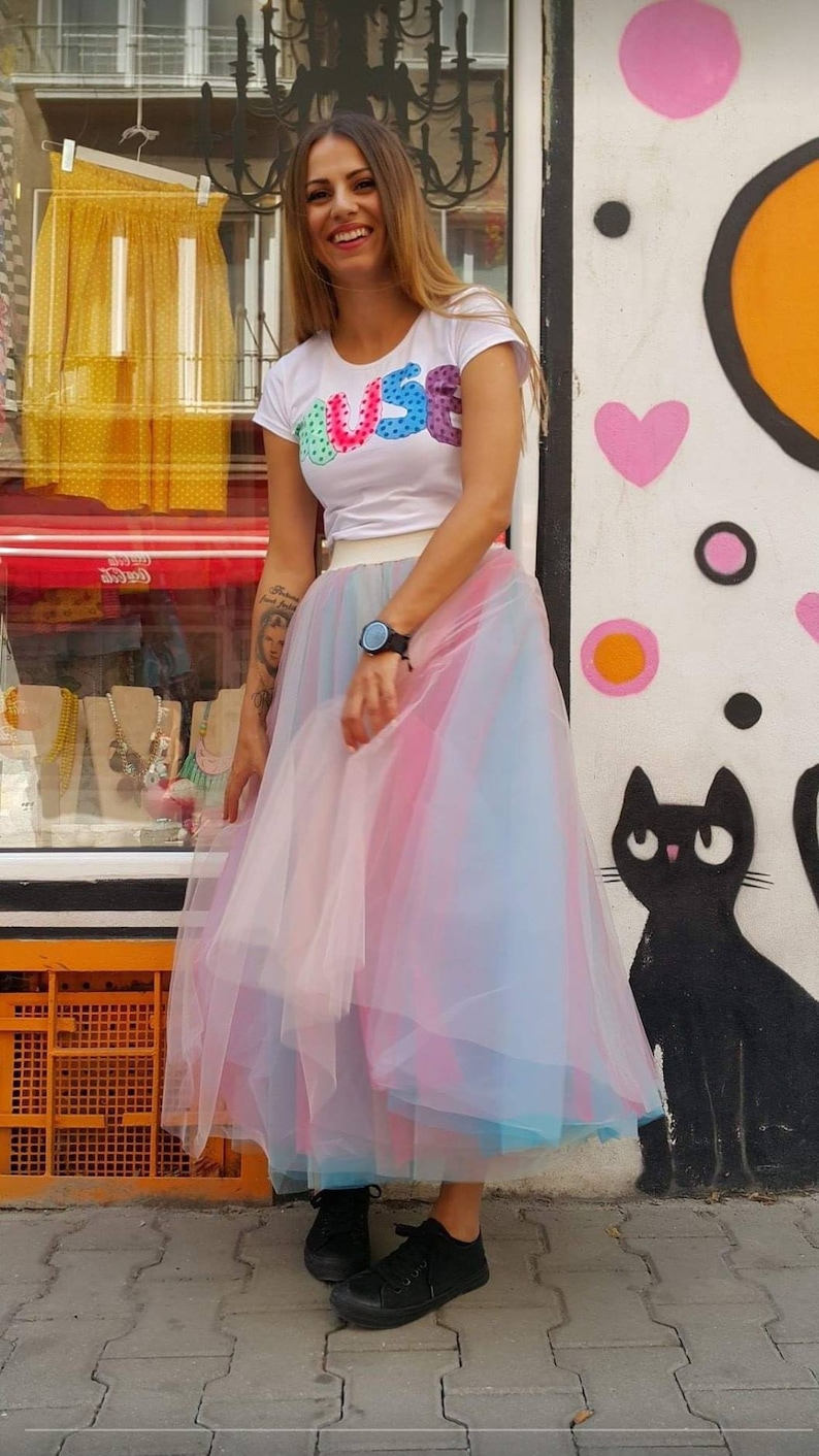 May include: A woman wearing a white t-shirt with the word "JUS&eacute;" in colourful letters and a rainbow tulle skirt. She is standing in front of a wall with colourful graffiti.