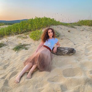以下が含まれることがあります： 茶色の巻き毛の女性が、グラフィックプリントの入った白いTシャツ、茶色のチュールスカート、茶色のベルトを着用して、砂浜に座っています。後ろには流木があります。