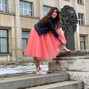 May include: A woman wearing a black leather jacket and a pink tulle skirt stands on a set of stone steps. She is putting on a pair of white heeled sandals.