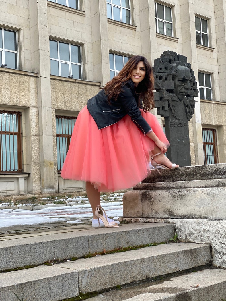 May include: A woman wearing a black leather jacket and a pink tulle skirt stands on a set of stone steps. She is putting on a pair of white heeled sandals.