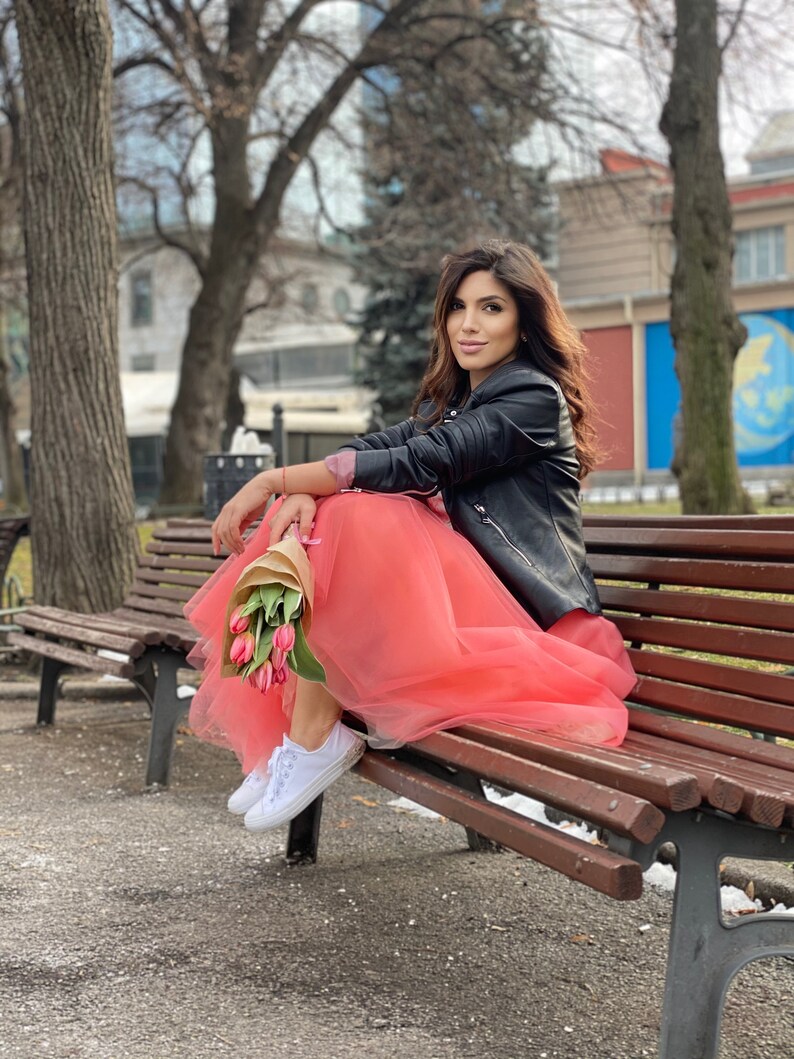 May include: A woman wearing a black leather jacket and a pink tulle skirt sits on a wooden bench. She is holding a bouquet of pink tulips.
