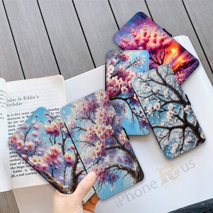 May include: Several phone cases with floral artwork. The cases display cherry blossom tree designs in various colours against blue and sunset backgrounds. The cases are open, showing the inside. The cases are on a white surface.