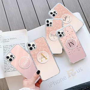 May include: Four pink and gold glitter iPhone cases with clear phone rings. The rings have different names and initials on them. The names are Ethan, India, Vick, and IN. The cases are all clear and have a glitter design.