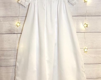 May include: White christening gown with a lace trim and pleats. The gown is hanging on a hanger against a woodgrain background.