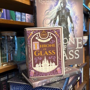 May include: A stack of books, including a hardcover edition of "Throne of Glass" by Sarah J. Maas. The book covers feature illustrations and text in shades of purple, gold, and white. The background shows a bookshelf filled with other books.