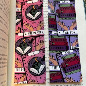 May include: Two pink and purple bookmarks with a tarot card design. The bookmarks feature a mountain, a crescent moon, a book, and a hand with long nails. The text on the bookmarks reads "The Reader" and "The Tower".