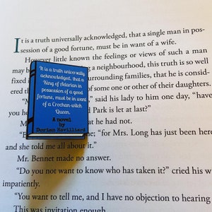 May include: A blue enamel pin featuring a book cover with the title "A Novel by Dorian Havilliard". The text on the book cover reads "It is a truth universally acknowledged, that a King of Adarlan in possession of a good fortune, must be in want of a Crochan-witch Queen."