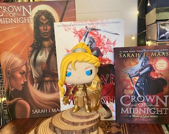 Puede incluir: Una figura Funko Pop dorada de una guerrera se encuentra sobre una exhibición de rodajas de madera, con tres portadas de libros detrás. Los libros se titulan "Crown of Midnight" de Sarah J. Maas. La figura tiene cabello rubio y ojos azules.