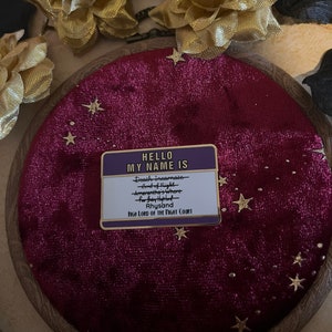 以下が含まれることがあります： 紫色の背景に金色の星が散りばめられた金色のピン。ピンには「HELLO MY NAME IS」というテキストと、「Death Incarnate - Lord of Night - Amarantha's Whore - Rhysand - HIGH LORD OF THE NIGHT COURT」というテキストが書かれたネームタグが付いています。