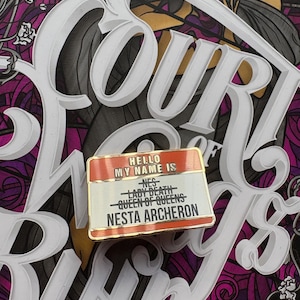 Op de afbeelding: Een rode en gouden emaille pin met de tekst "Hello my name is" en een lijst met namen: "Nes", "Lady Death", "Queen of Queens" en "Nesta Archeron".