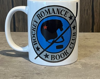 May include: White ceramic mug with a black and gray graphic of a hockey helmet and stick with the text "Hockey Romance Book Club" in a circle.