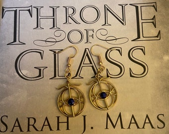 Puede incluir: Un par de pendientes de metal dorado con un diseño de alambre circular y una cuenta azul oscuro en el centro, suspendidos de ganchos. Los pendientes están colocados sobre el texto de la portada del libro Throne of Glass de Sarah J. Maas.