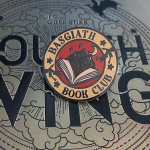 Pode incluir: Um broche de esmalte dourado e vermelho com um livro preto, uma lua crescente e estrelas. O broche diz "Basgiath Book Club" em letras pretas.