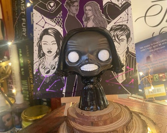 Puede incluir: Una figura pop en blanco y negro de un personaje con una túnica negra con capucha y una cara de calavera blanca. La figura está de pie sobre una plataforma de madera con tres niveles. El fondo es una portada de libro con un diseño morado y negro y texto que dice "A Court of Mist and Fury" y "A Court of Thorns and Roses".