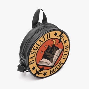 Pode incluir: Mochila redonda preta com uma alça superior e uma alça de ombro ajustável. A frente apresenta um design circular com o texto "BASGIATH BOOK CLUB" em torno de um gráfico de uma montanha, livro aberto, lua e estrelas.