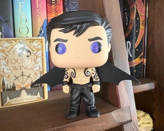 May include: A vinyl collectible figure with black hair, purple eyes, and black wings. The figure has black arm bands and black pants, with black swirl tattoos on the chest and shoulders. The figure stands on a flat surface with a neutral skin tone.