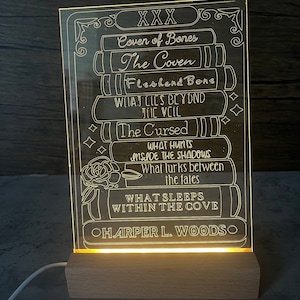 Könnte beinhalten: Ein beleuchtetes Acrylschild mit einem Stapel Bücher und dem Text "XXX", "Cover of Bones", "The Coven", "Fleshend Bone", "What Lies Beyond The Veil", "The Cursed", "What Hunts Inside The Shadows", "What Lurks Between The Fates", "What Sleeps Within The Cove" und "Harper L. Woods".