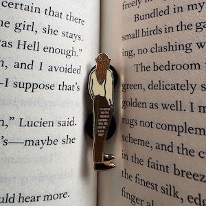 May include: A metal enamel pin featuring a figure with long reddish-brown hair, wearing a cream-colored top and brown trousers. The figure is standing with their back turned, and the text on the trousers reads: IF I OFFER YOU THE MOON ON A STRING, WILL YOU GIVE ME A KISS, TOO?