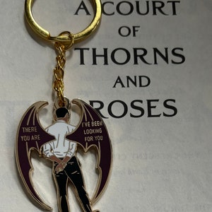 Puede incluir: Llavero dorado con un dije que presenta una figura con alas de murciélago. El dije tiene un contorno dorado, una camisa blanca, pantalones negros y las frases "THERE YOU ARE" y "I'VE BEEN LOOKING FOR YOU".