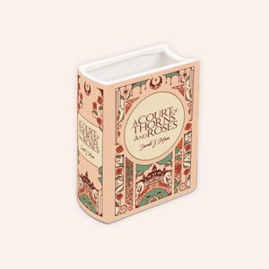 May include: A decorative ceramic book-shaped vase with the title "A Court of Thorns and Roses" in a vintage style. The vase is peach-colored with floral and architectural design elements in red, green, and cream. The interior is white.