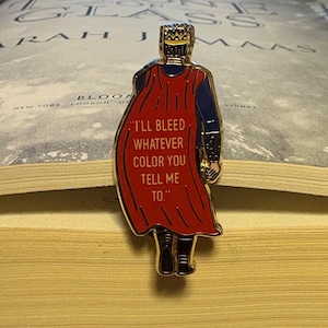 Peut inclure: &Eacute;pingle en &eacute;mail dor&eacute;e repr&eacute;sentant une figurine couronn&eacute;e avec une cape rouge. La cape porte l'inscription "I'LL BLEED WHATEVER COLOR YOU TELL ME TO." L'&eacute;pingle est pos&eacute;e sur les pages ouvertes d'un livre.
