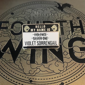 May include: A gold-colored metal pin with a black background and white text. The pin features a dragon design and the text "Hello My Name Is - Violence - Silver One - Violet Sorrengail".