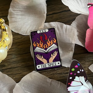 May include: A purple tarot card with the number XXIV and the text "THE SPICE" on it. The card features a book with flames and two hands reaching out from the book. There are red chilli peppers on top of the book.