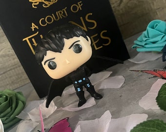 Puede incluir: Una figura de vinilo pop negra y azul de un personaje con cabello negro y alas. La figura está de pie sobre una superficie blanca con un libro titulado "A Court of Thorns and Roses" en el fondo.