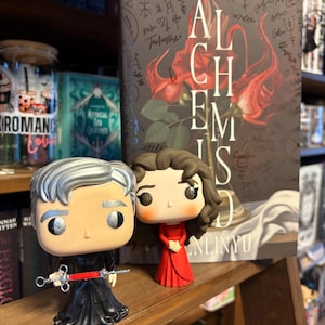 May include: Two Funko Pop figures stand before a book titled "ALCHEMIST". One figure has silver hair and a syringe, the other wears a red dress. The book cover features red roses and the title in large white letters.