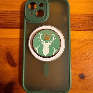 Puede incluir: Una funda de teléfono verde y transparente con un emblema circular que presenta un ciervo blanco con cuernos y una corona dorada sobre un fondo verde. La funda tiene un borde verde, sobre una superficie de madera.