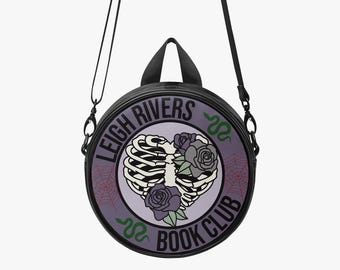 Puede incluir: Bolso bandolera redondo negro con un diseño de caja torácica en forma de corazón, rosas moradas y el texto "LEIGH RIVERS BOOK CLUB". Tiene un asa superior y una correa ajustable.