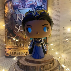 Puede incluir: Una figura de vinilo pop azul y dorada de una mujer que lleva una corona y una túnica azul y dorada. La figura tiene ojos azules y piel morena. La figura está de pie sobre una plataforma de madera. El fondo es un libro con el título "King's Scourge" y una cadena de luces.