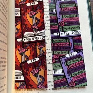 May include: Two tarot cards with a purple and pink color scheme. The card on the left is labeled "XXII" and the card on the right is labeled "XXX". Both cards have a sword and roses on them. The card on the right has the text "The Tropes" and lists "Enemies to Lovers", "Love Triangle", "Fated Mates", "Reverse Harem", "One Bed", "Forbidden Love", "Gage", and "Forced Proximity".