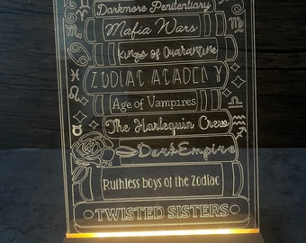 May include: A rectangular, illuminated acrylic book stack night light. The light features etched titles like "Darkmore Penitentiary" and "Mafia Wars." The base is a light-colored wooden block. A white cord extends from the base.