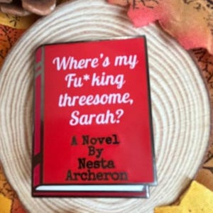 May include: A red book-shaped pin with the title "Where's my Fu*king threesome, Sarah?" and the author's name, Nesta Archeron. The pin is set against a neutral background with autumn-themed decorations.