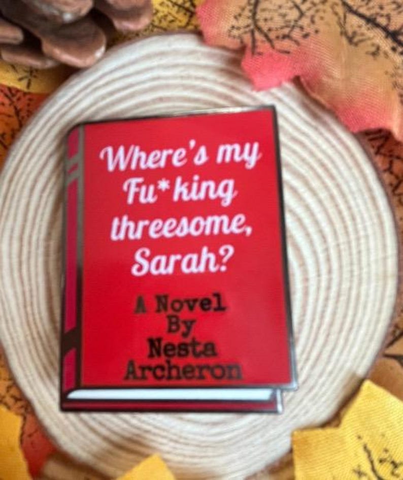 May include: A red book-shaped pin with the title "Where's my Fu*king threesome, Sarah?" and the author's name, Nesta Archeron. The pin is set against a neutral background with autumn-themed decorations.