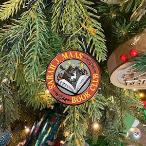 Peut inclure: Une décoration de Noël ronde avec le texte "Sarah J. Maas Book Club". L'ornement présente une scène de montagne avec un loup et un livre ouvert. La décoration est suspendue à un sapin de Noël.