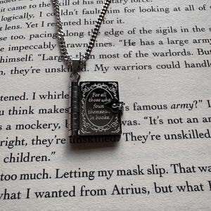 Può includere: Collana color argento con un piccolo ciondolo a forma di libro incernierato. Il ciondolo nero reca l'iscrizione "For all those who found themselves in books". La collana è esposta su una pagina stampata.