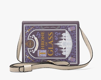 Może przedstawiać: Torba na ramię z motywem inspirowanym książką "Throne of Glass". Torba ma fioletową okładkę ze złotymi literami i ilustracją białego zamku. Posiada kremowy pasek i złote okucia.