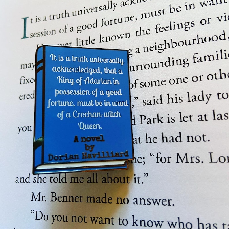May include: A blue enamel pin shaped like a book with the title "A novel by Dorian Havilliard" and the text "It is a truth universally acknowledged, that a King of Adarlan in possession of a good fortune, must be in want of a Crochan-witch Queen."