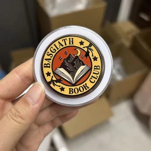 Puede incluir: Un soporte circular para teléfono con un diseño de Basgiath Book Club. El gráfico incluye un libro abierto, montañas oscuras, una luna creciente y la silueta de un dragón volador sobre un fondo naranja, dentro de un borde amarillo con texto negro.