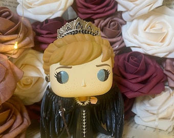 May include: A black and gold pop vinyl figure of a woman wearing a black dress and a crown. She has wings and is holding a staff. The figure is surrounded by artificial flowers in shades of white, pink, and burgundy.