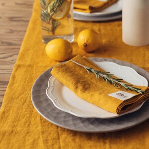Pode incluir: Um corredor de mesa de linho amarelo com um prato branco e uma toalha de linho amarela dobrada. Um ramo de alecrim é colocado sobre a toalha. Há dois limões e um copo de água com limões e alecrim na mesa.