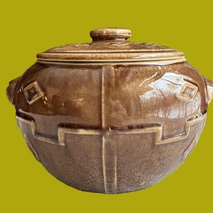May include: A brown ceramic crock with a lid, featuring a geometric design. The crock has two handles on the sides and a glossy finish.